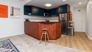 Cocina compacta con armarios azul oscuro, un taburete de madera, un refrigerador de acero inoxidable y un microondas.