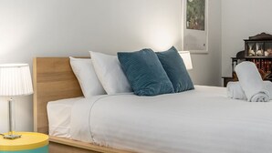 Una cama bien hecha con ropa blanca y almohadas turquesa, una lámpara de noche y una mesita redonda con tapa amarilla.