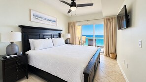 A hotel room with a large bed, a ceiling fan, a TV mounted on the wall, and a view of the ocean through the window. - Emerald Isle 1202, Gulf Front, Beach Chair Service Included (Pensacola Beach)