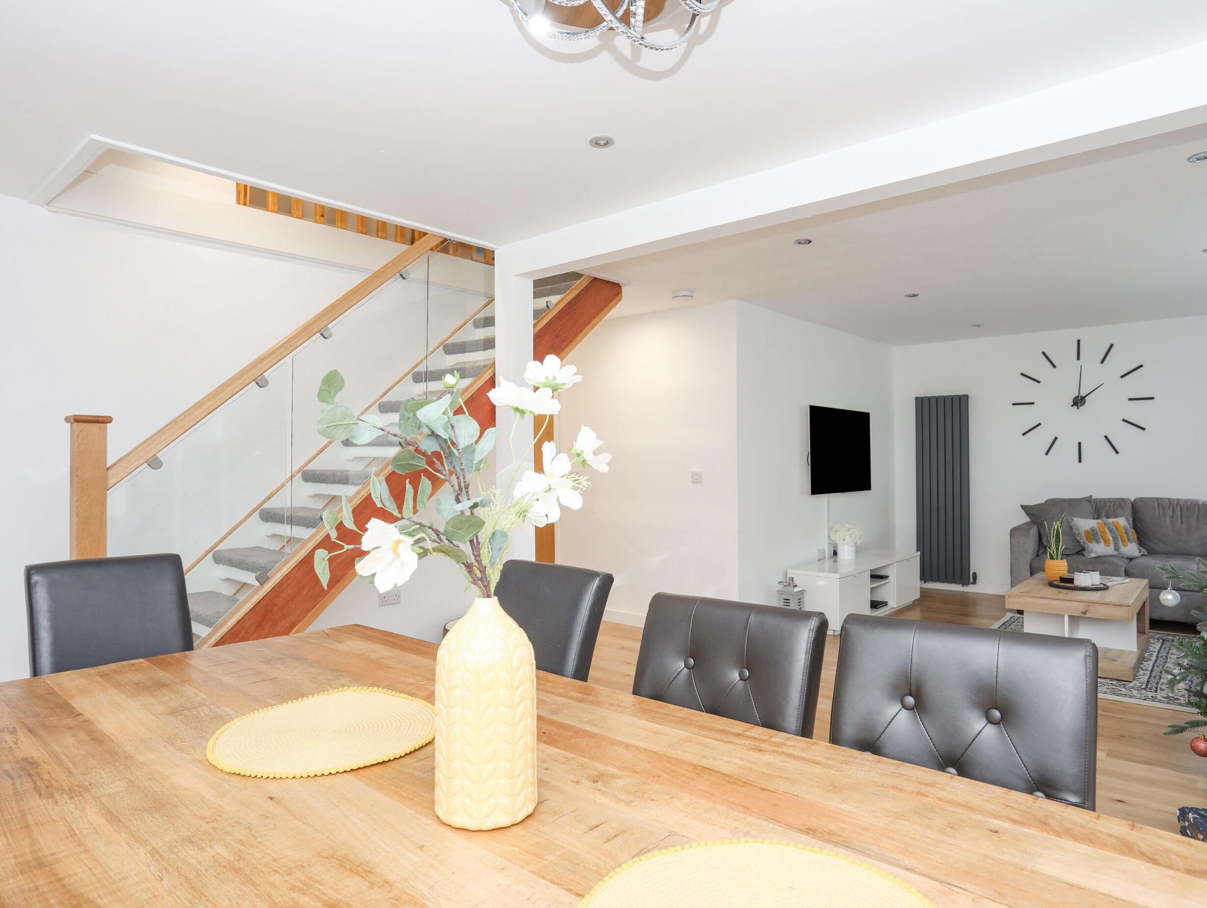 A modern living room with a dining area, featuring a wooden dining table, black leather chairs, a staircase with wooden railings, a flat-screen TV, and a large wall clock.