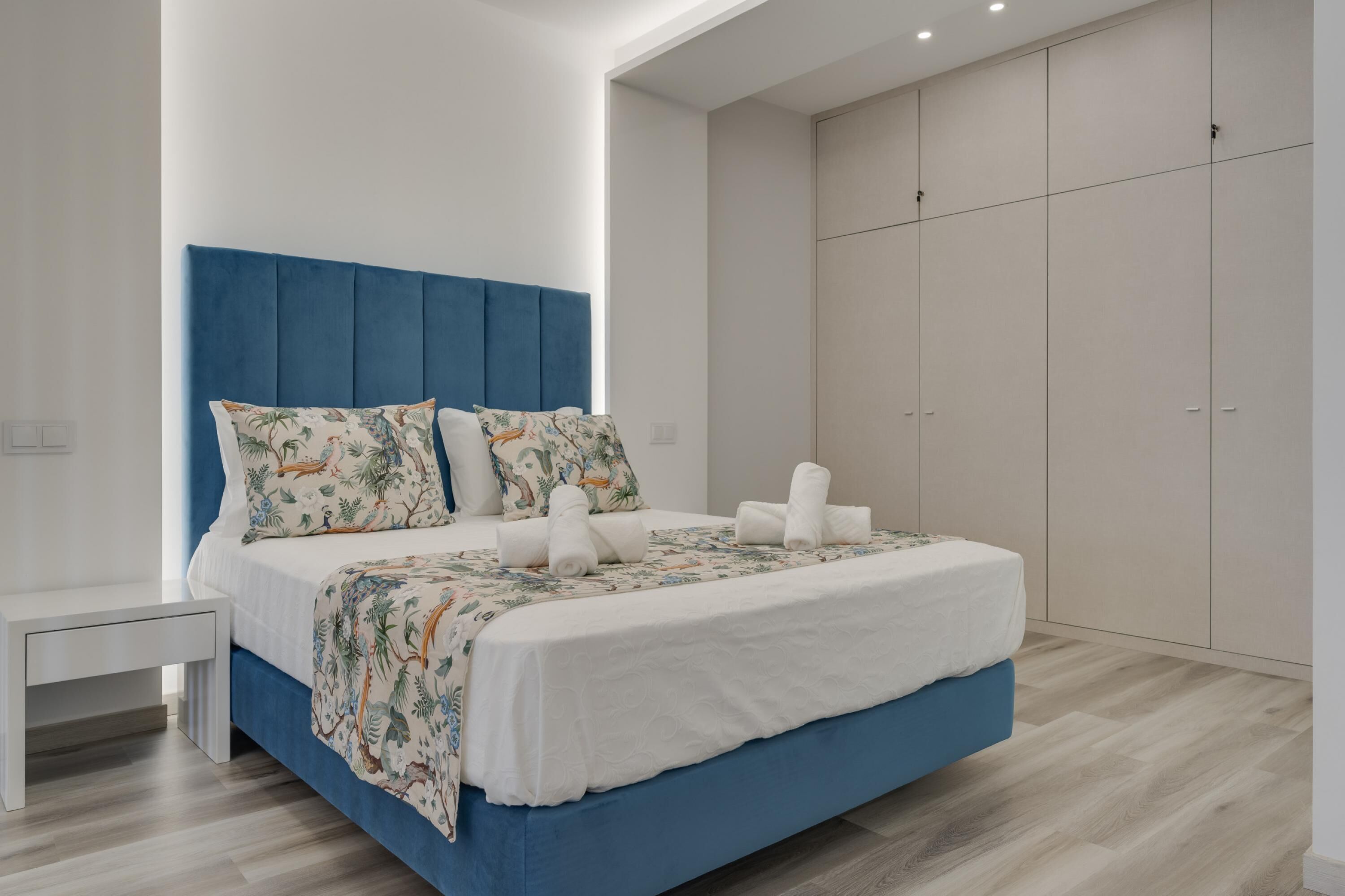 A modern bedroom with a blue upholstered headboard, a white bedspread with a tropical print, and neatly folded towels on the bed.