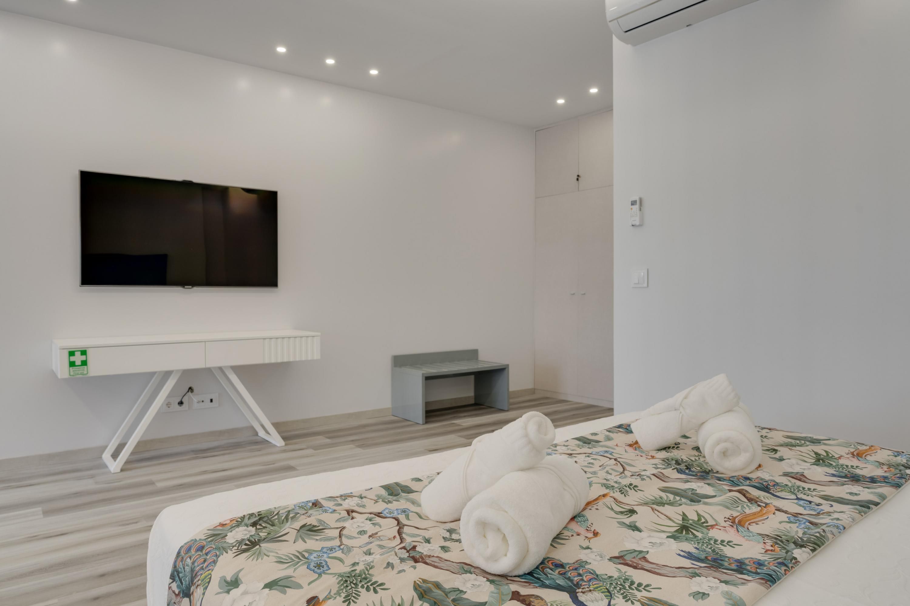 A modern hotel room with a large bed, a flat-screen TV mounted on the wall, a small desk, and a grey bench.