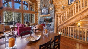 A cozy log cabin interior with a dining area, stone fireplace, and a wooden staircase.