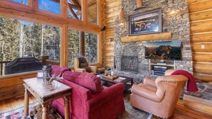 A log cabin interior with a stone fireplace, a television mounted above it, and a large window overlooking a forest.