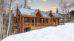 A log cabin with a stone foundation, wooden exterior, and metal roofing, surrounded by snow and trees.