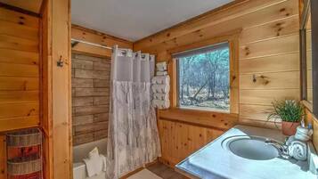 A wooden cabin bathroom with a shower curtain, a window with a view of trees, and a sink with a towel.