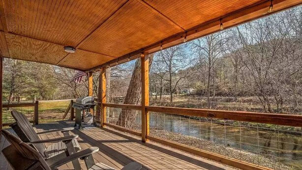 A covered outdoor area with wooden flooring, a grill, and a view of a river and trees. - Pet Friendly Creekside Cabin + Hot Tub + Fire Pit (Mineral Buff)