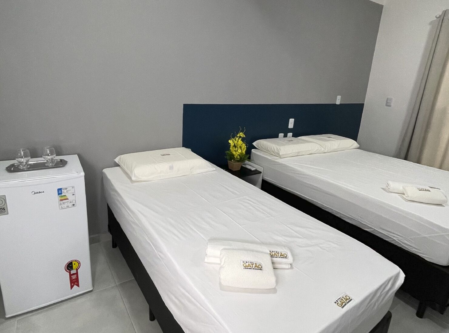 Habitación de hotel con dos camas individuales, un minibar y una mesita con una planta en maceta.