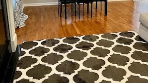 Uma área de refeições com mesa e cadeiras de madeira, um tapete estampado e uma janela com um painel decorativo.