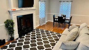 Uma sala de estar com lareira, uma TV montada acima dela, um espaço de refeições com mesa e cadeiras, e um tapete estampado.