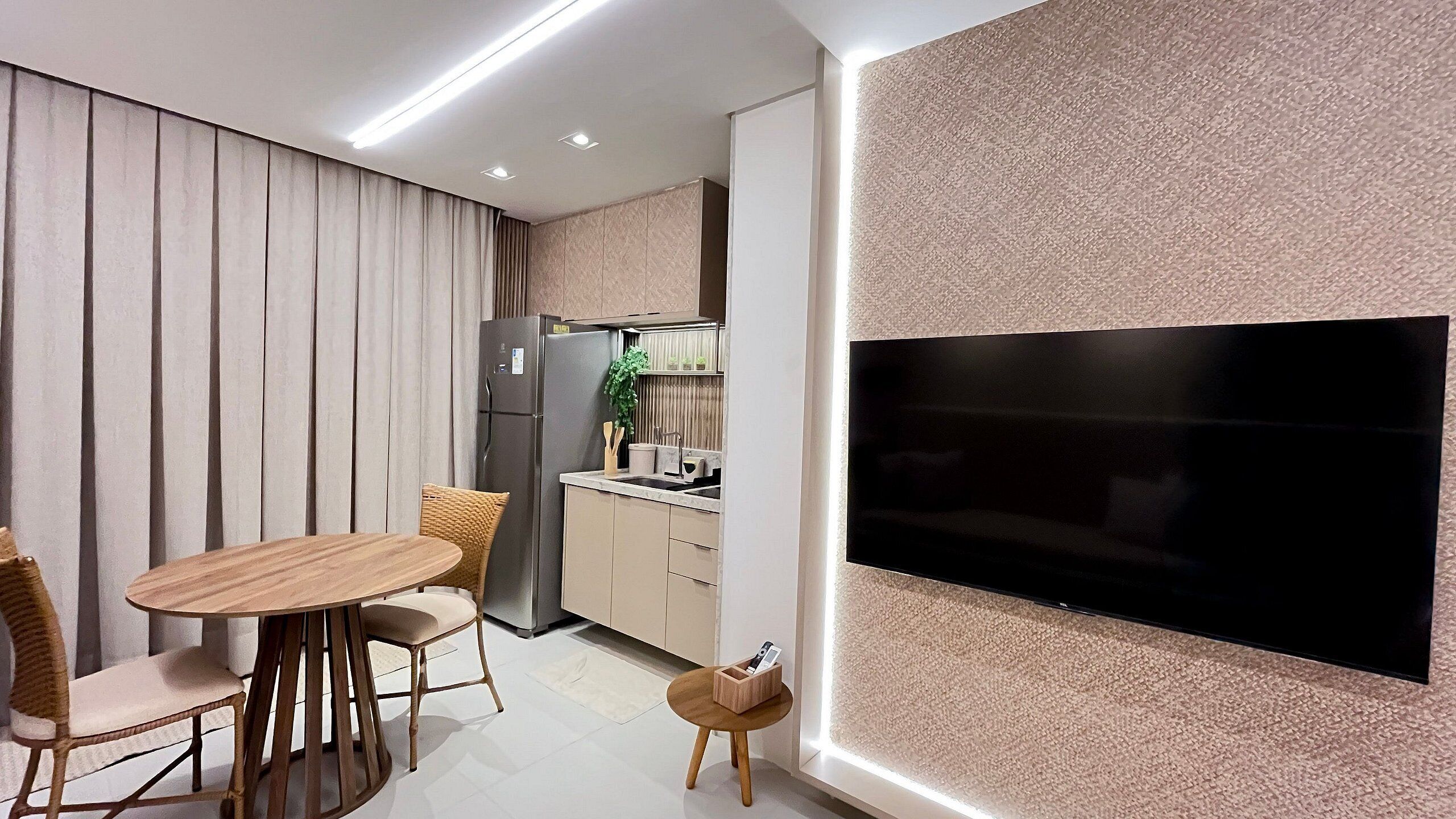 A modern kitchen with a built-in refrigerator, a dining area with a round wooden table and chairs, and a large flat-screen TV mounted on the wall.