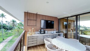 Ein Balkon mit Esstisch und Stühlen, eine Küchenzeile mit Fernseher und Blick auf Palmen.