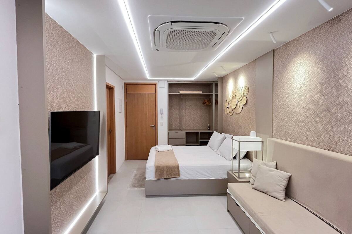 A modern hotel room with a flat-screen TV, a bed with a beige blanket, a sofa, a bedside table, and a walk-in closet.