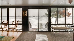 Une pièce dotée de grandes fenêtres donnant sur une plage enneigée, avec des chaises en bois, un banc et un tableau encadré accroché au mur.
