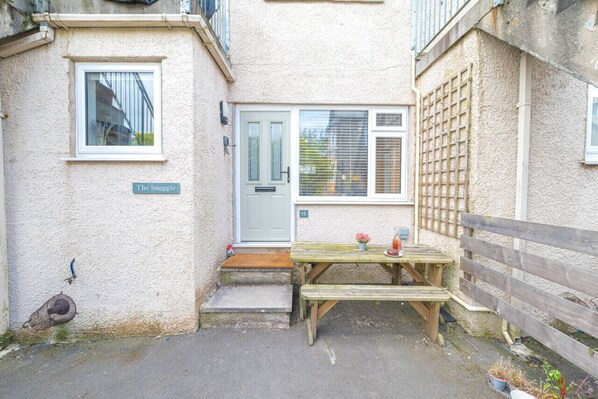 A small, cozy courtyard with a wooden bench, a door labeled 'The Snuggle', and a small table with a plant and a bottle. - Great getaway for couples at this central Windermere ground floor flat with parking (Windermere)