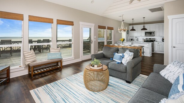 A spacious living area with a large window overlooking a deck, a grey sofa, a wooden bench, and a kitchen in the background.