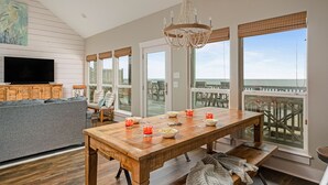 A spacious living room with a wooden dining table set for a meal, a grey sofa, and a television on the wall. Large windows offer a view of the beach and pier.