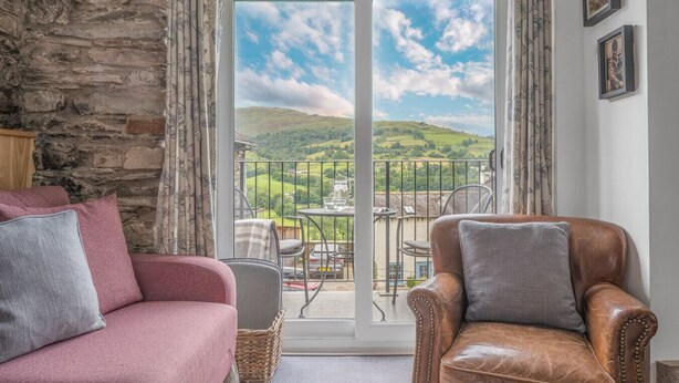 A cozy living room with a stone wall, a pink sofa, a brown leather armchair, and a view of green hills through a glass door. - Romantic getaway, little two bed, two bath barn conversion with amazing views & parking (Ambleside)