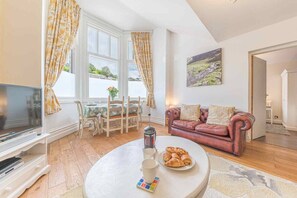 Ein gemütliches Wohnzimmer mit Fernseher, einem runden Couchtisch mit Gebäck, einem roten Ledersofa und einem Bild an der Wand.