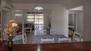 A dining area with a wooden table and chairs, a vase of flowers, and a view into a living room with a sofa and a window with blinds. - Holiday apartment on the quiet (Vrbnik)