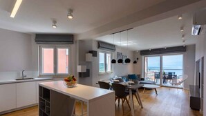 Cocina moderna con isla central, comedor con sillas y un amplio ventanal con vistas al mar.