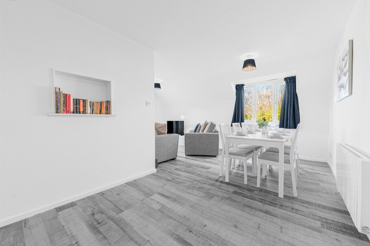 A modern, minimalist living room with a dining area, featuring a bookshelf, grey sofas, and a dining table with chairs.