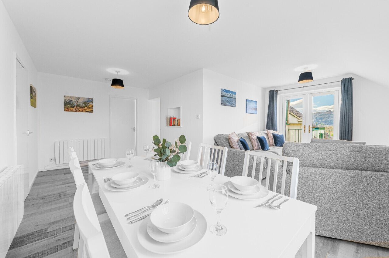 A modern dining area with a white table set for four, a grey sofa, and a window with curtains.