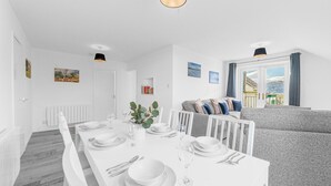 A modern dining area with a white table set for four, a grey sofa, and a window with curtains. - Two minute walk from the World of Beatrix Potter, apartment with parking (Bowness-on-Windermere)