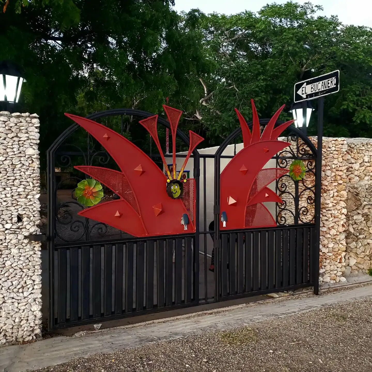 Uma entrada fechada com um portão de metal decorativo com formas abstratas vermelhas e uma placa de rua com a inscrição “FL BUCANERO”.