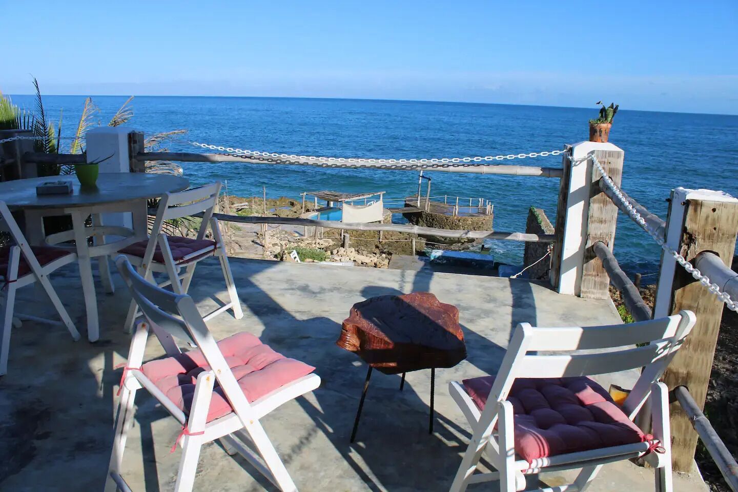 Un espacio al aire libre con sillas blancas y una mesa, con vista a una costa rocosa y al mar.