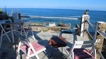 Un espacio al aire libre con sillas blancas y una mesa, con vista a una costa rocosa y al mar.