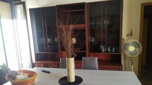 Ein Essbereich mit Tisch, Stühlen, einer Vase mit Trockenpflanzen, einem Korb voller Früchte und einem Ventilator.