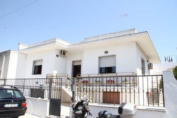 Un edificio moderno blanco con entrada protegida por una reja, una escalera y un coche aparcado.