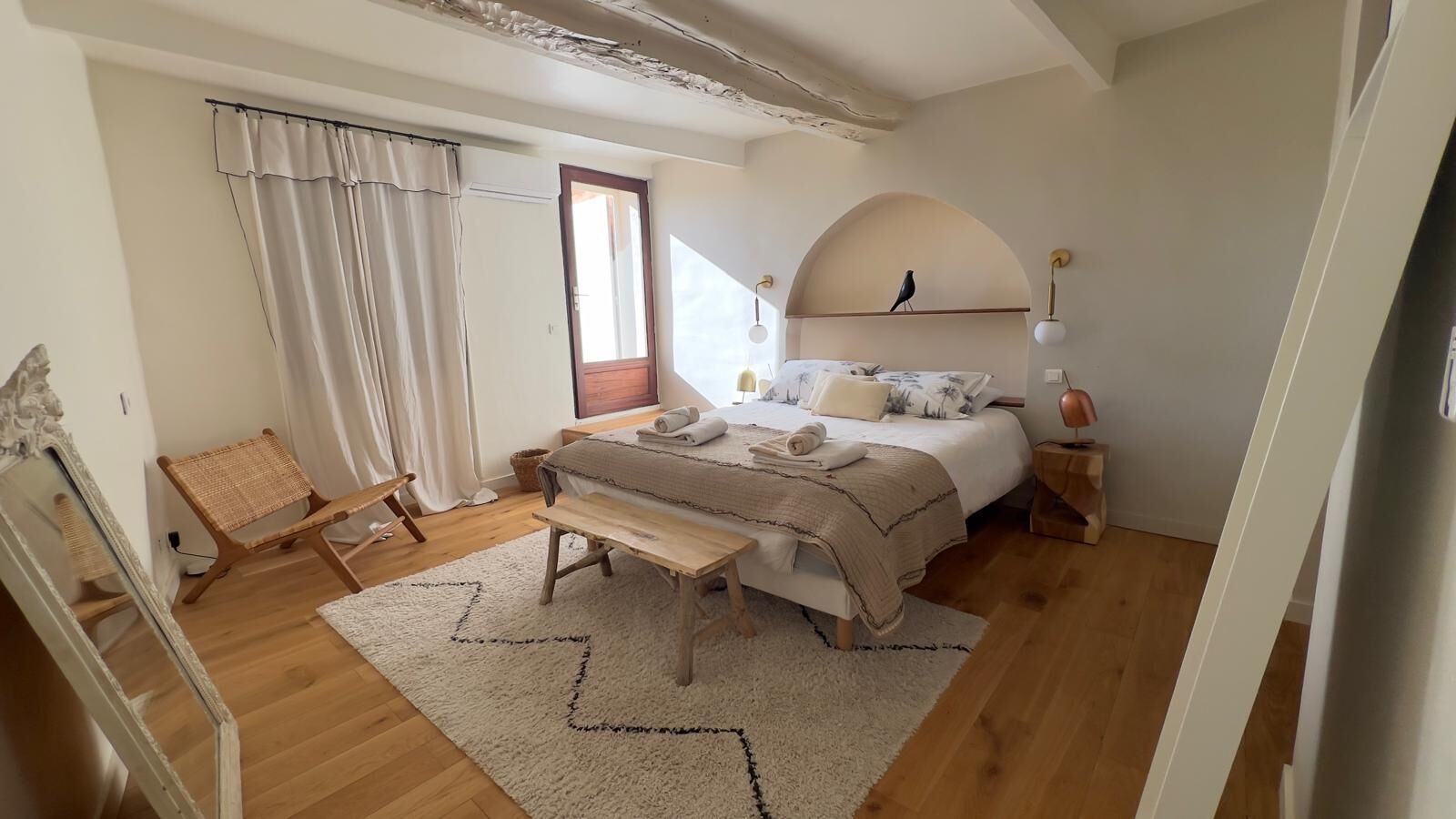 Een slaapkamer met een bed, een bankje, een stoel, een spiegel, een raam met gordijnen en een lamp.