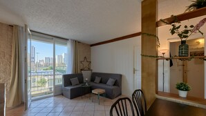 Ein Wohnzimmer mit grauem Sofa, einem kleinen Tisch und einem Esstisch mit Stühlen. Ein großes Fenster bietet Blick auf Gebäude und einen Balkon.