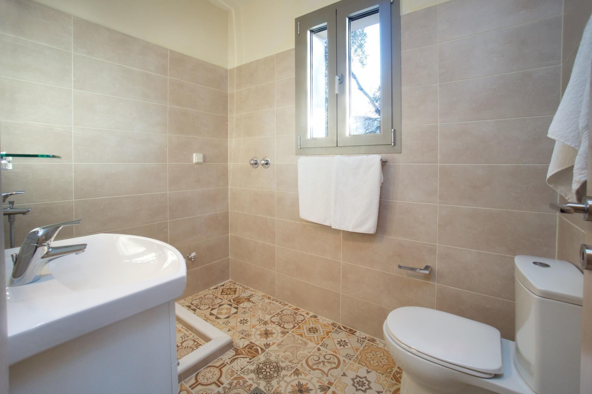 A bathroom with beige tiles, a white sink, a toilet, and a window above the sink.