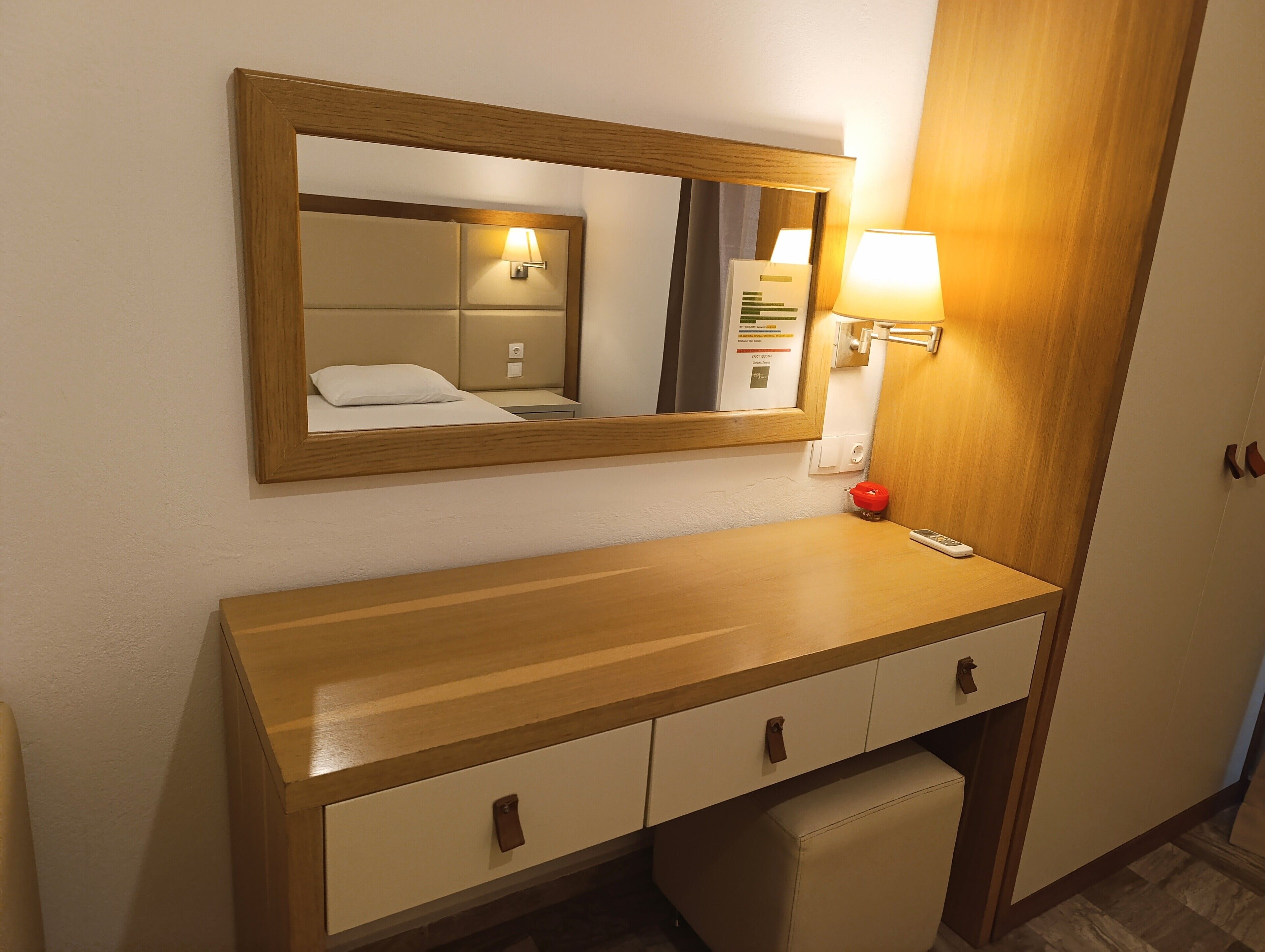 A hotel room with a wooden vanity, a mirror, a bedside lamp, a red object on the counter, and a small ottoman.