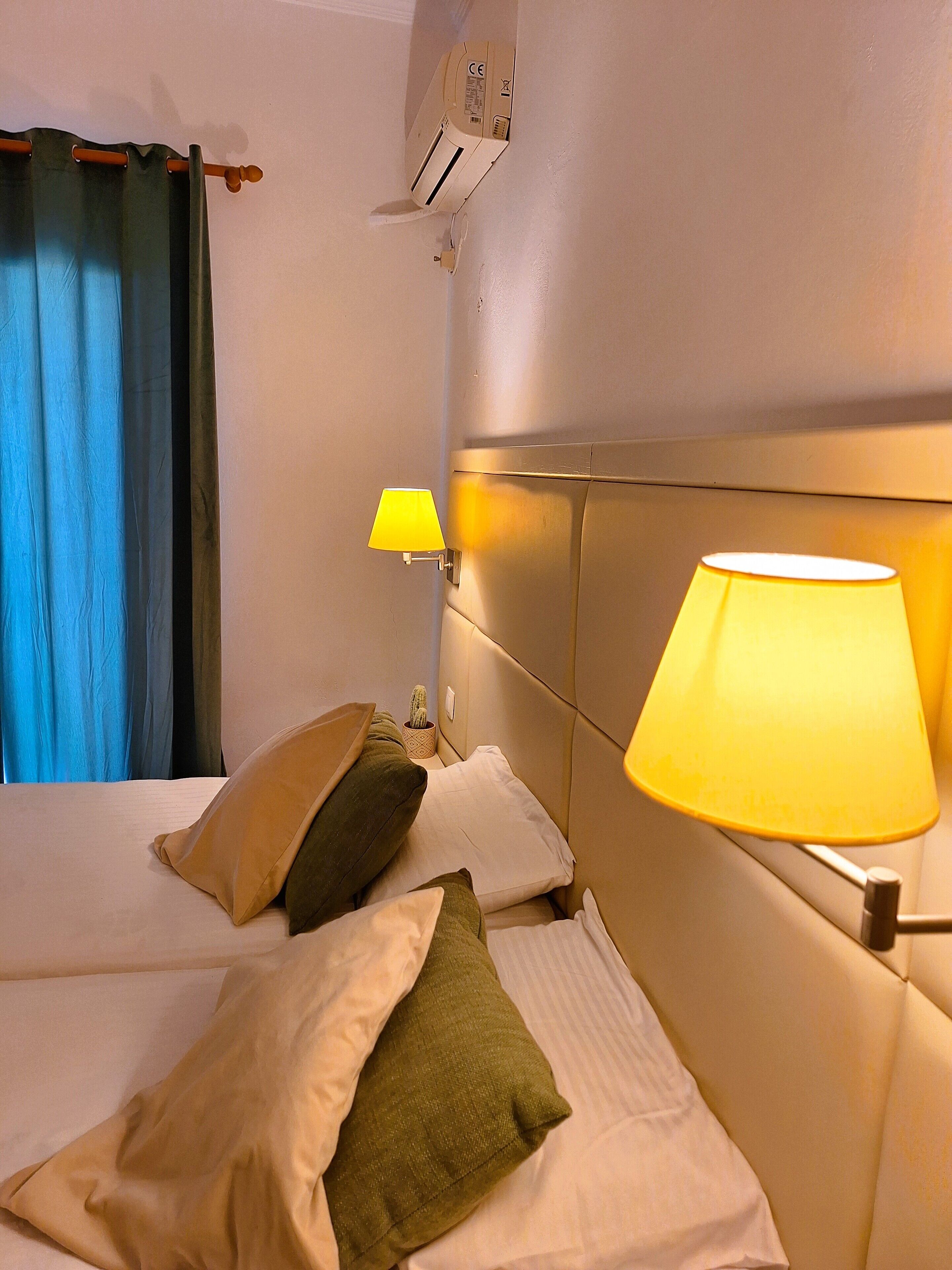 A hotel room with a bed, pillows, a lamp, an air conditioning unit, and a window with curtains.