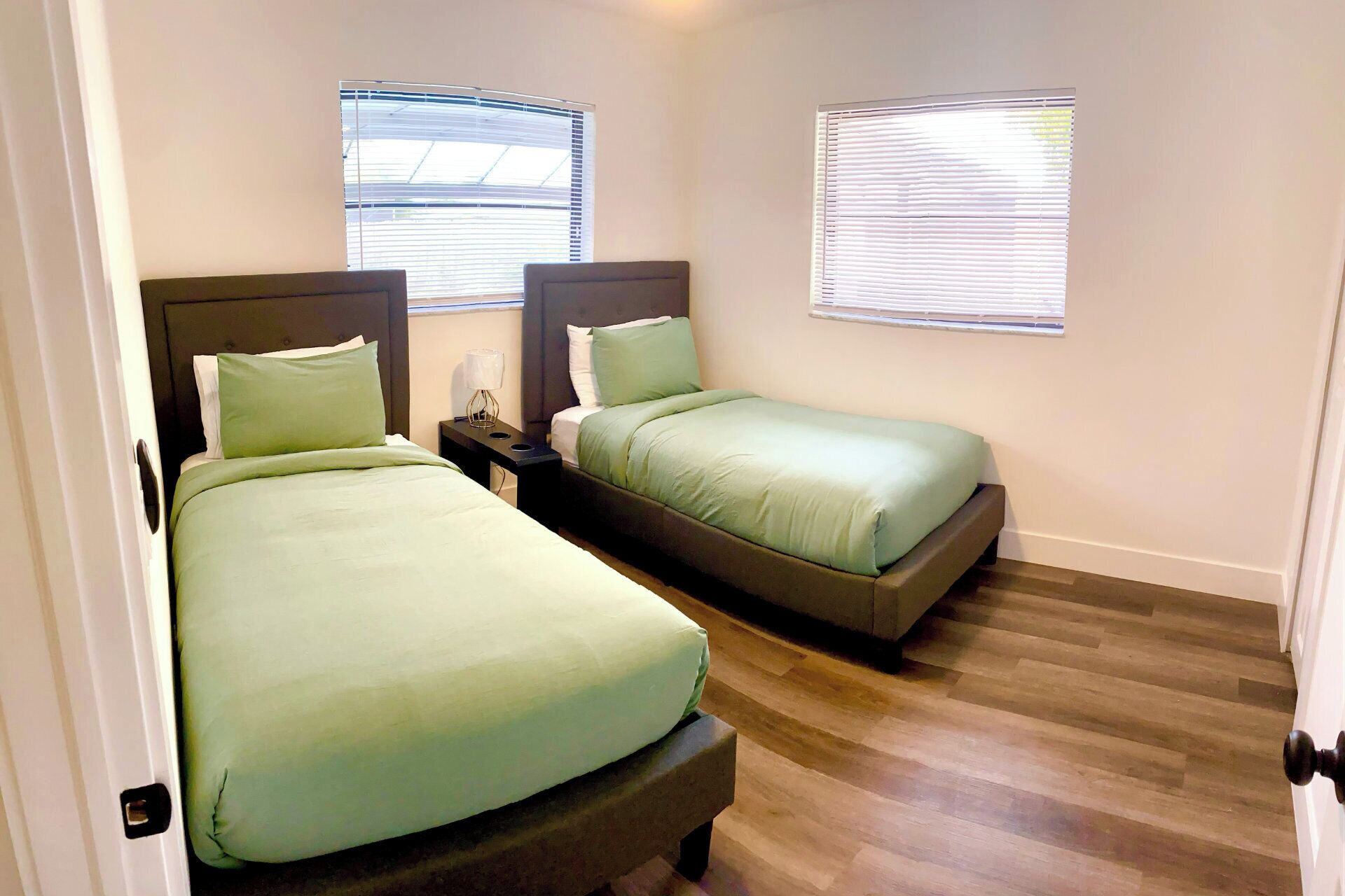 A bedroom with two beds, a nightstand with a lamp, and two windows with blinds.