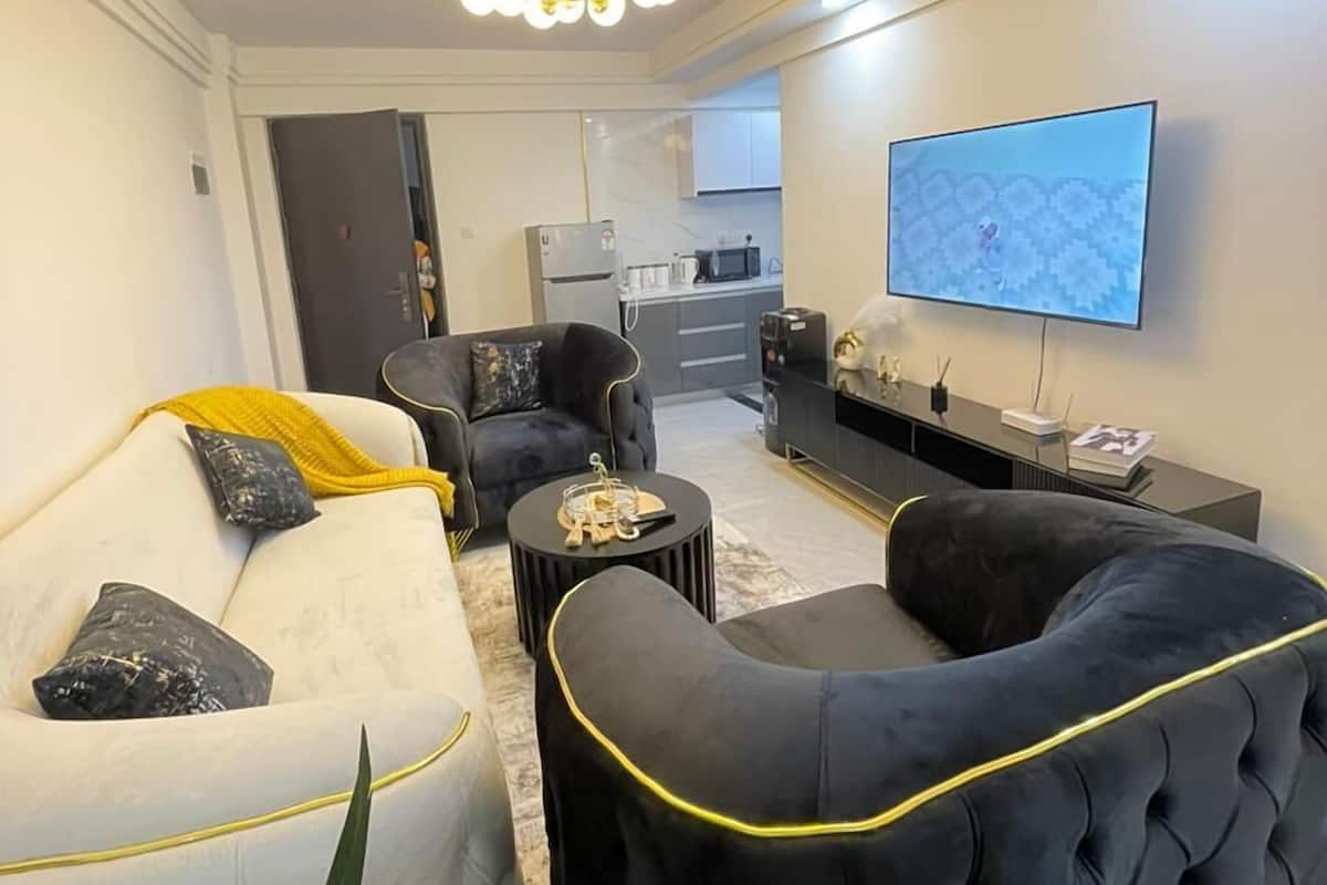 A modern living room with a sofa, armchair, and coffee table. A flat-screen TV is mounted on the wall.