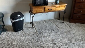 A room with a wooden table, a black trash bin, a microwave, and a dresser.