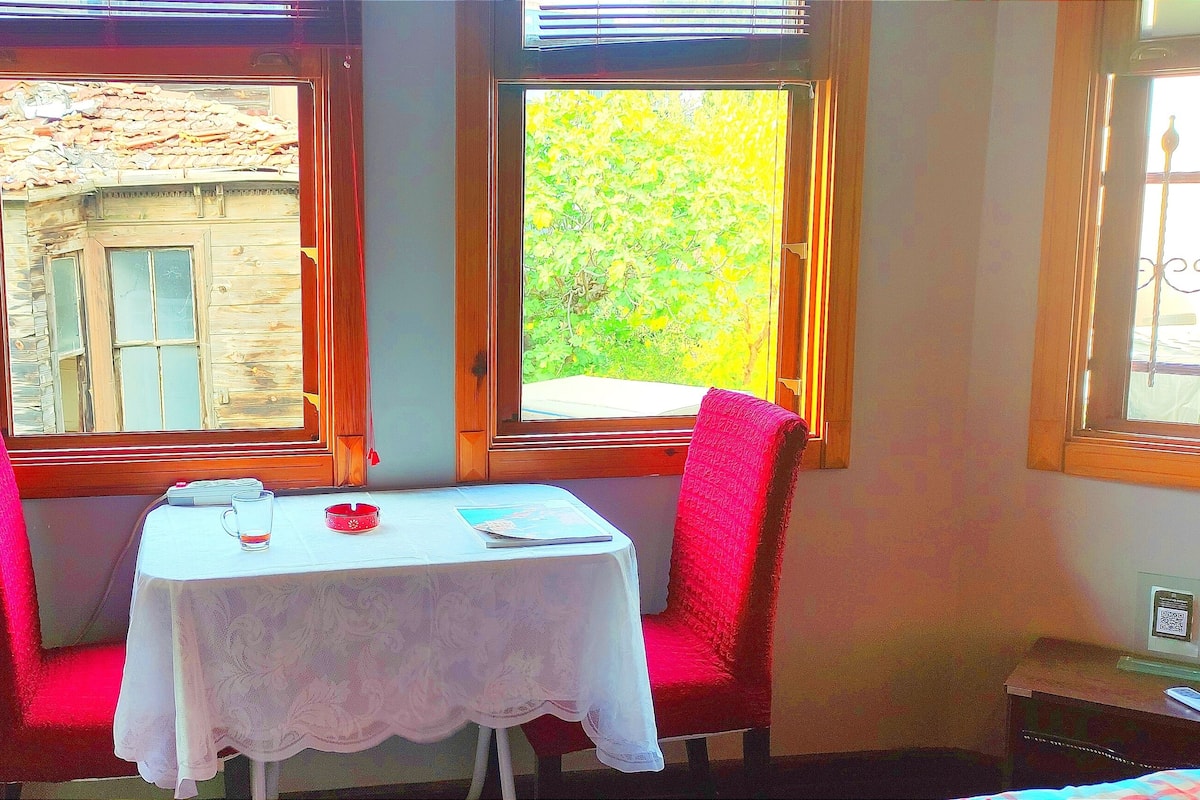 A corner room with a table and two chairs, a window with a view of a building and greenery, and a small bedside table with a phone and a QR code.