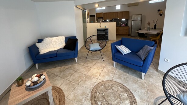A modern living room with two blue sofas, a small table with decorative items, and a kitchen area in the background.