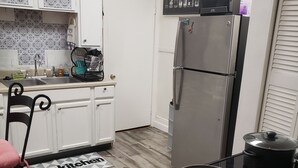 Una cocina con armarios blancos, un refrigerador de acero inoxidable, un microondas y una estufa. Hay una silla negra con un cojín rosa, un escurreplatos y una felpudo con el texto 'I love my kitchen'.