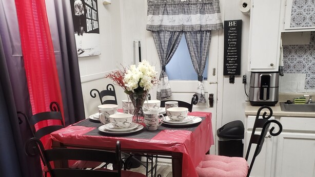 Un comedor con una mesa dispuesta para cuatro personas, rodeada de sillas de metal con cojines rosa. Un jarrón con flores blancas está sobre la mesa, y una cafetera en la encimera.