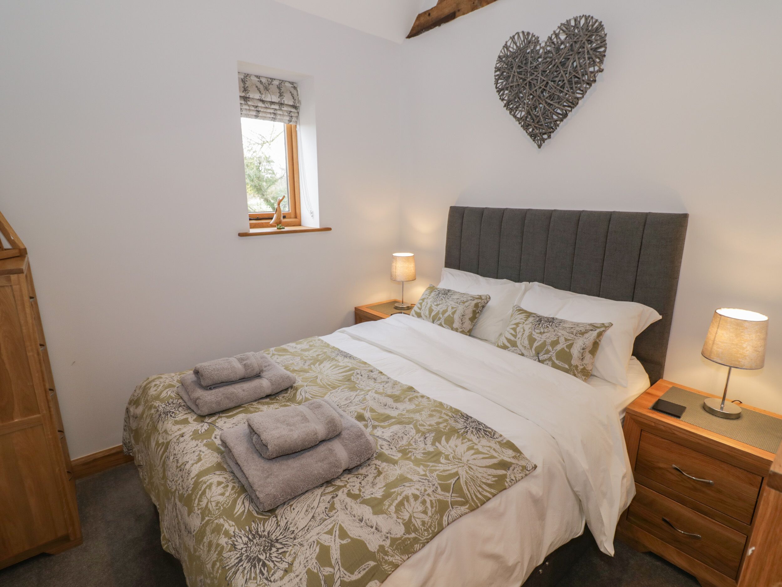 Una cama bien hecha con un edredón estampado con flores, dos lámparas de noche y una decoración de corazón en la pared.