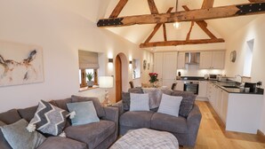 A spacious living room with a large grey sofa set, a small ottoman, and a dining area with a table and chairs. The kitchen is visible in the background with white cabinets and a wooden ceiling. - The Parlour (Droitwich)