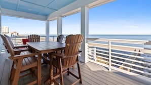A wooden deck with a table and chairs, overlooking a beach with a white fence and buildings in the distance. - Enjoy AMAZING VIEWS at almost new spacious oceanfront unit, huge deck, elevator (Carolina Beach)