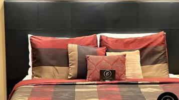 Un lit avec une tête de lit noire, une couette rouge et beige, et plusieurs coussins aux motifs variés.