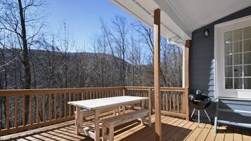 Un patio in legno con tavolo e panchina, un barbecue e vista su alberi e colline.